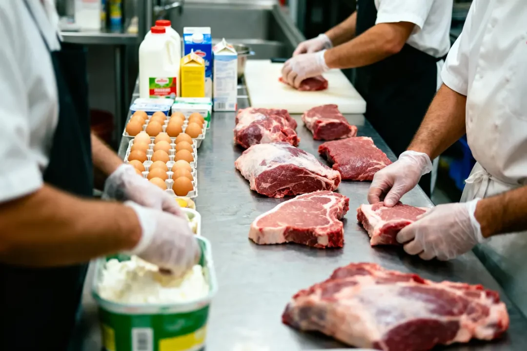 Cuisiniers préparant viande et œufs sur table inox, ingrédients variés en arrière-plan, ambiance de cuisine professionnelle.